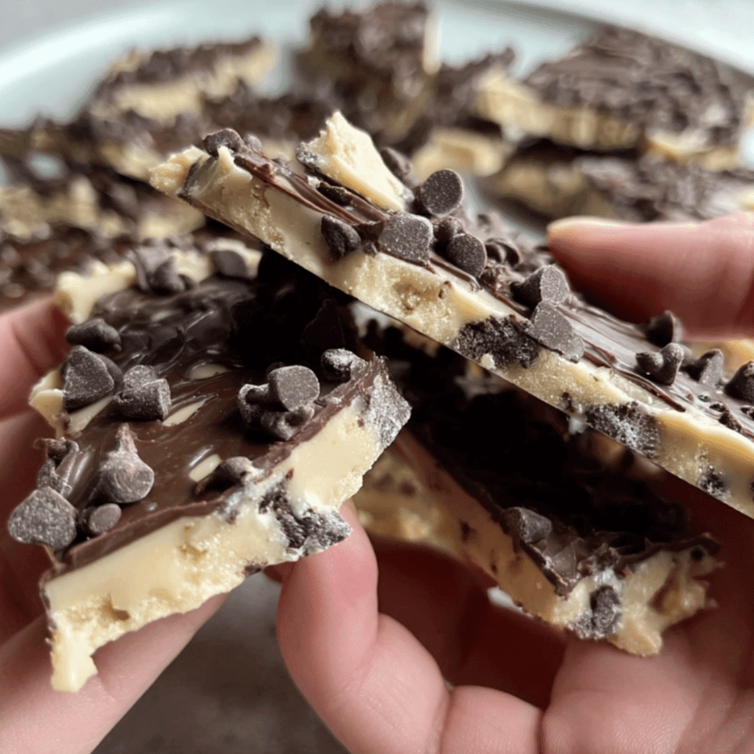 Hands holding a broken piece of chocolate-covered cookie dough bark showing the creamy center and chocolate chips