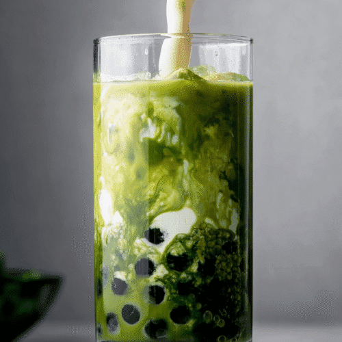 Creamy milk swirling into iced matcha tea with tapioca pearls in a clear glass.
