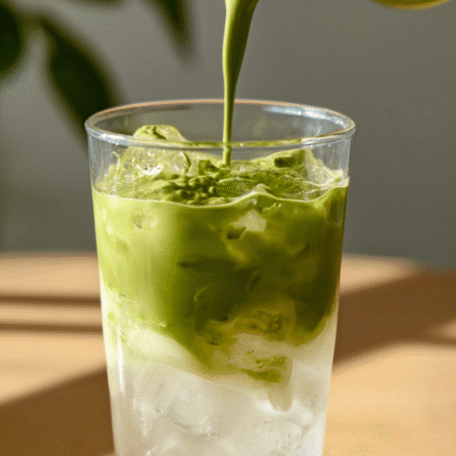 Whipped matcha being poured over iced coconut water in a glass on a wooden table.