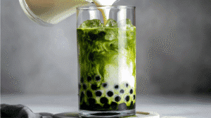 A tall glass of matcha bubble tea with dark tapioca pearls at the bottom and creamy milk swirling into vibrant green tea.